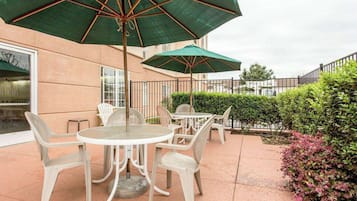 Un patio exterior con mesas redondas y sillas blancas, cubierto por grandes sombrillas verdes.