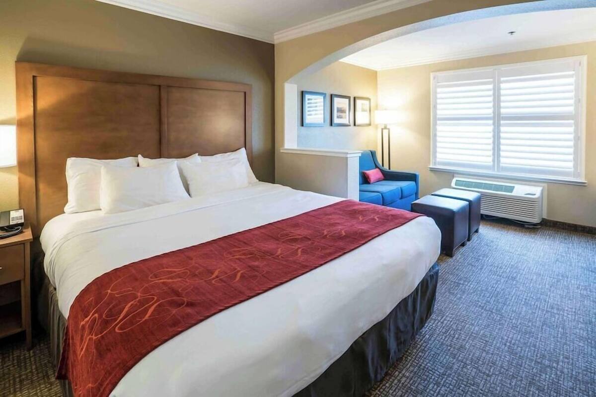 A hotel room with a large bed, a desk with a lamp, a chair, and a window with blinds.