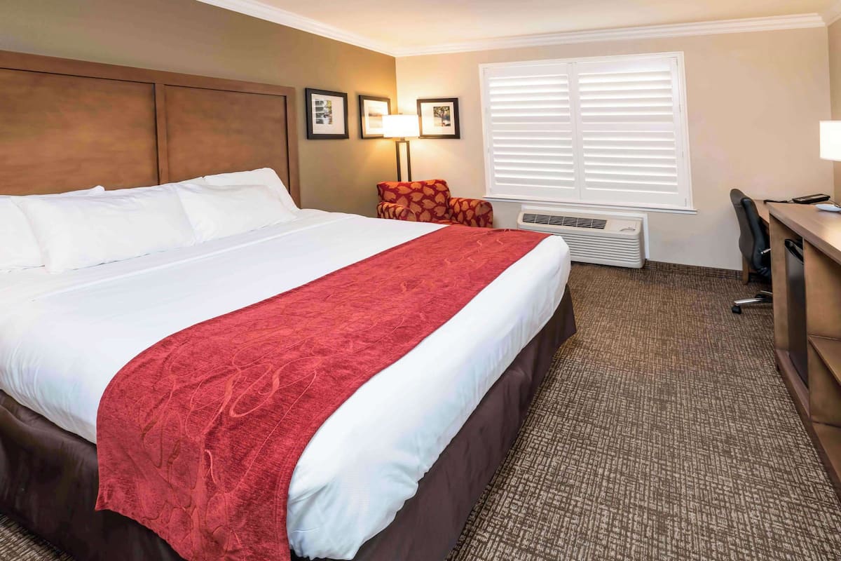 A hotel room with a large bed, a desk, a chair, a lamp, and a window with blinds.