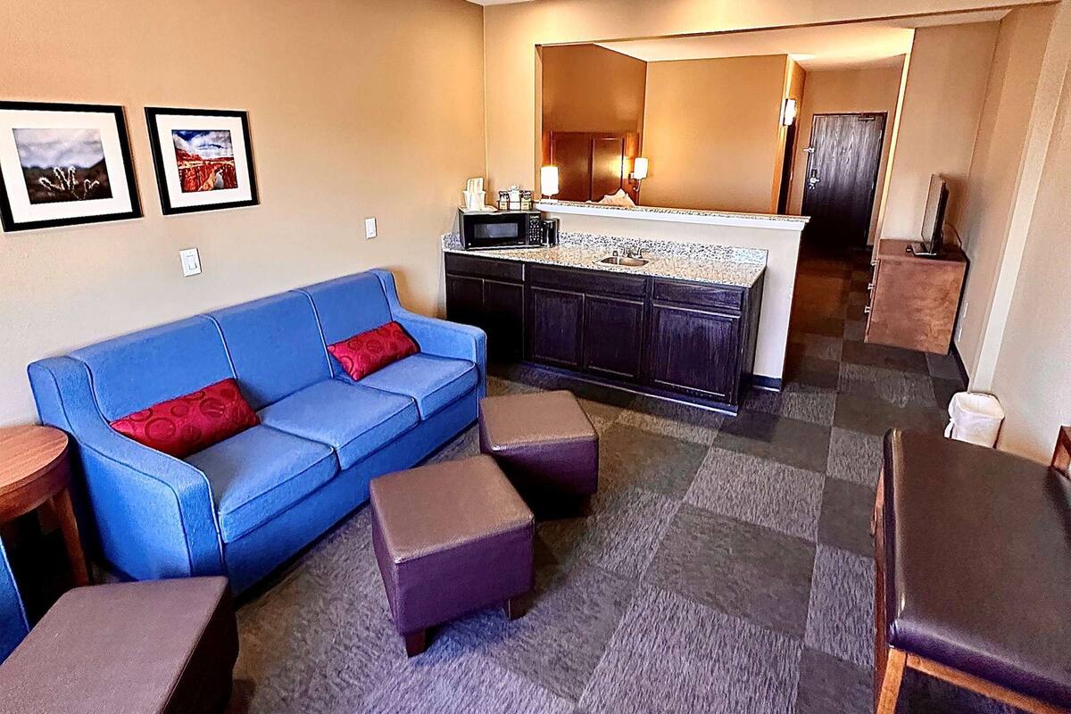 Una sala de estar con un sofá azul, dos otomanas, una mesa de centro y una cocina con microondas y fregadero.