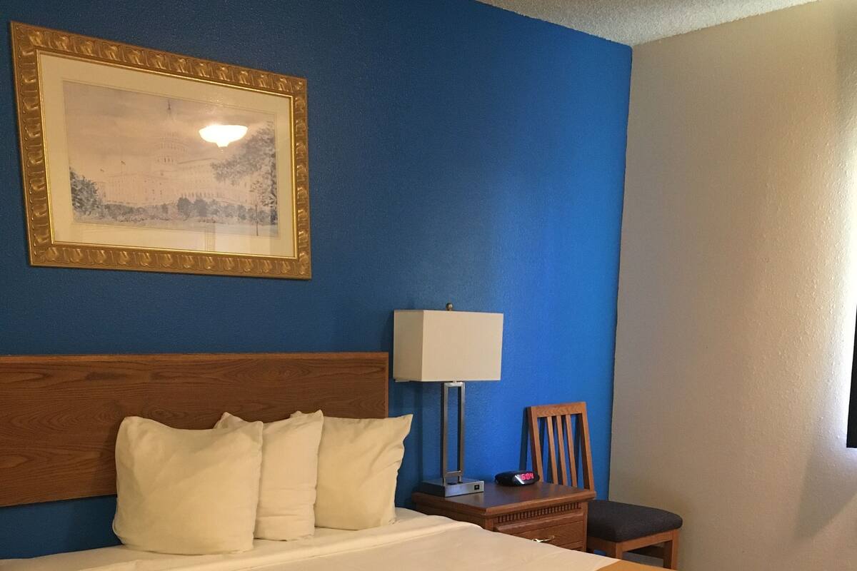 A hotel room with a bed, a nightstand, a lamp, a clock, and a framed picture on the wall.