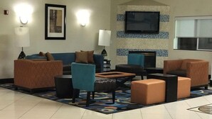 Un vestíbulo de hotel con un sofá azul, un sillón verde azulado y un reposapiés estampado. Hay un televisor de pantalla plana montado en la pared y una chimenea.