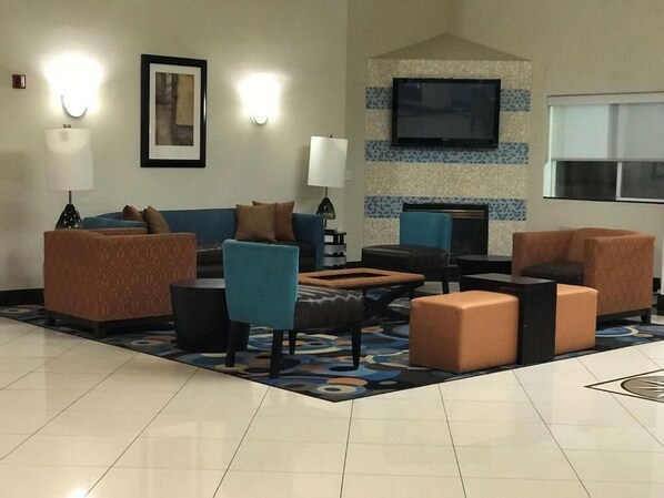 A hotel lobby with a blue sofa, a teal armchair, and a patterned ottoman. There is a flat-screen TV mounted on the wall and a fireplace.