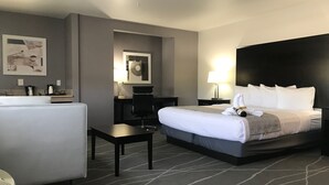 Habitación de hotel con cama, escritorio, silla, cafetera, lámpara y un cuadro en la pared.