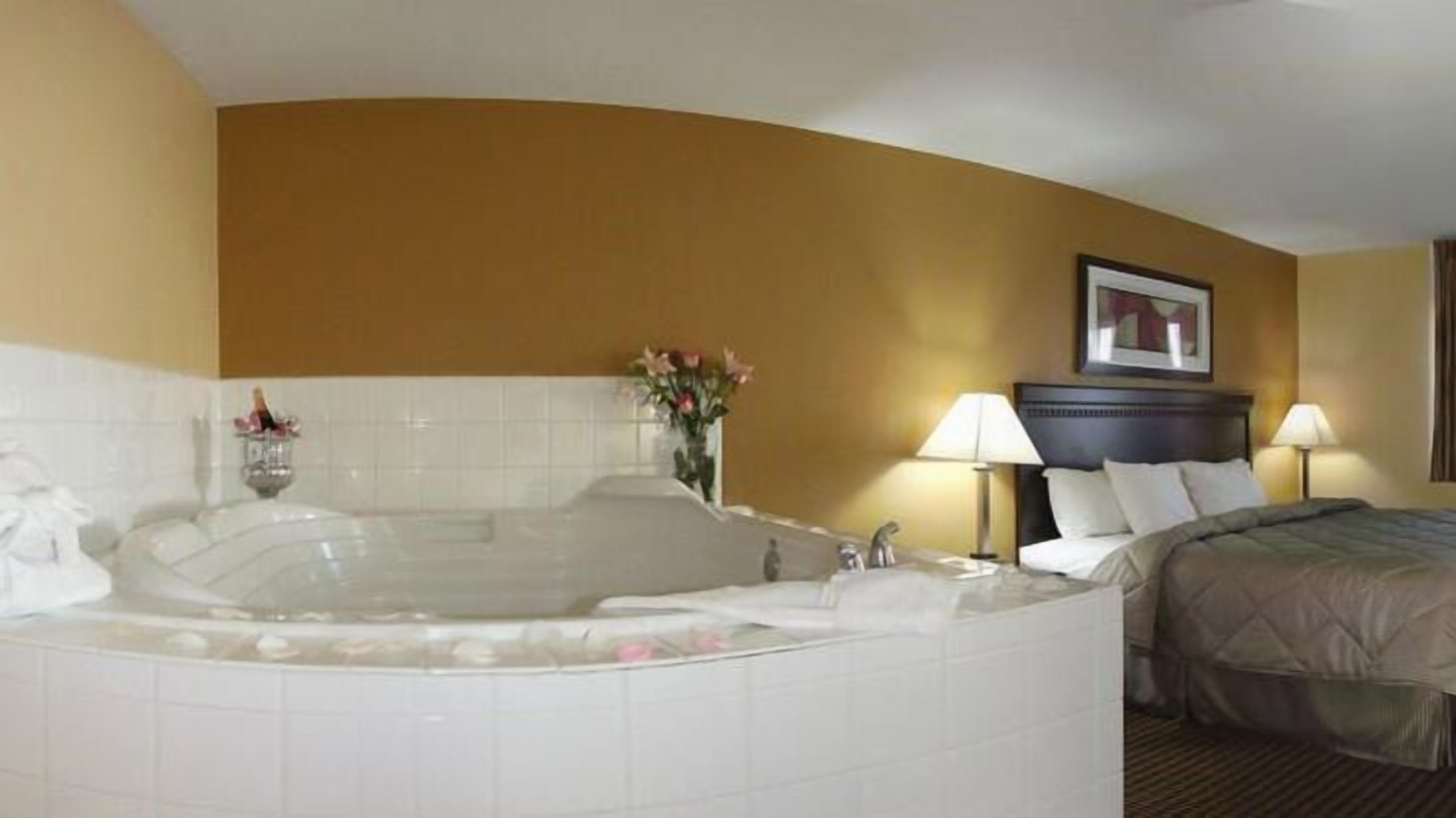 A hotel room with a large bathtub, a bed with a headboard, two bedside lamps, and a framed picture on the wall.