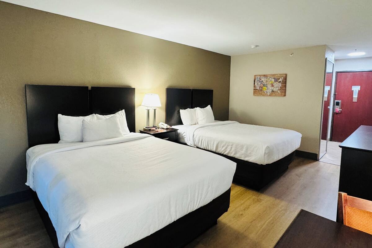 Habitación de hotel con dos camas, una mesita de noche con lámpara, un cuadro en la pared y una puerta con marco rojo.
