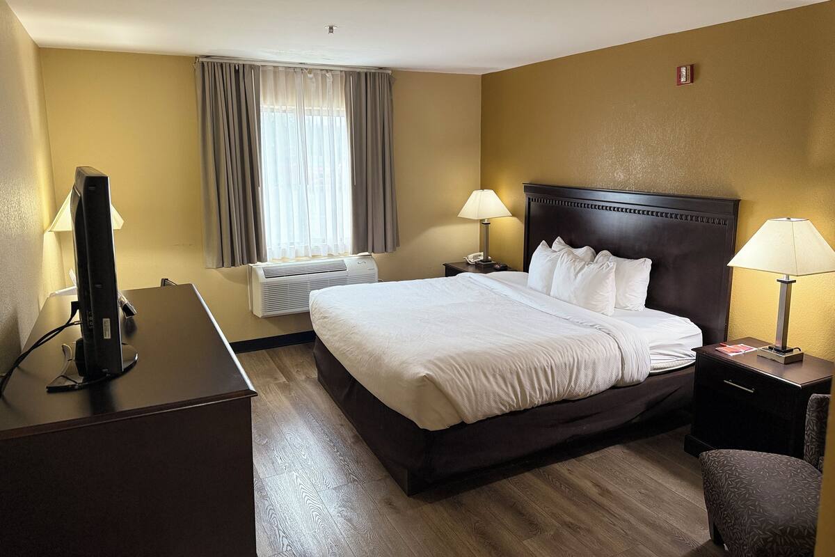 Una habitación de hotel con una cama, dos lámparas de noche, un escritorio con un ordenador, una ventana con cortinas y un aire acondicionado instalado en la pared.