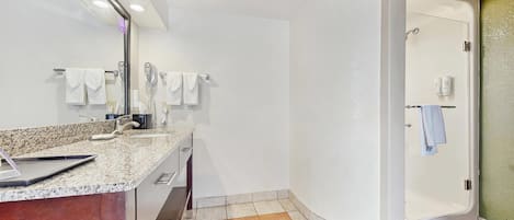 Un baño moderno con encimera de granito, espejo, secador de pelo y zona de ducha.