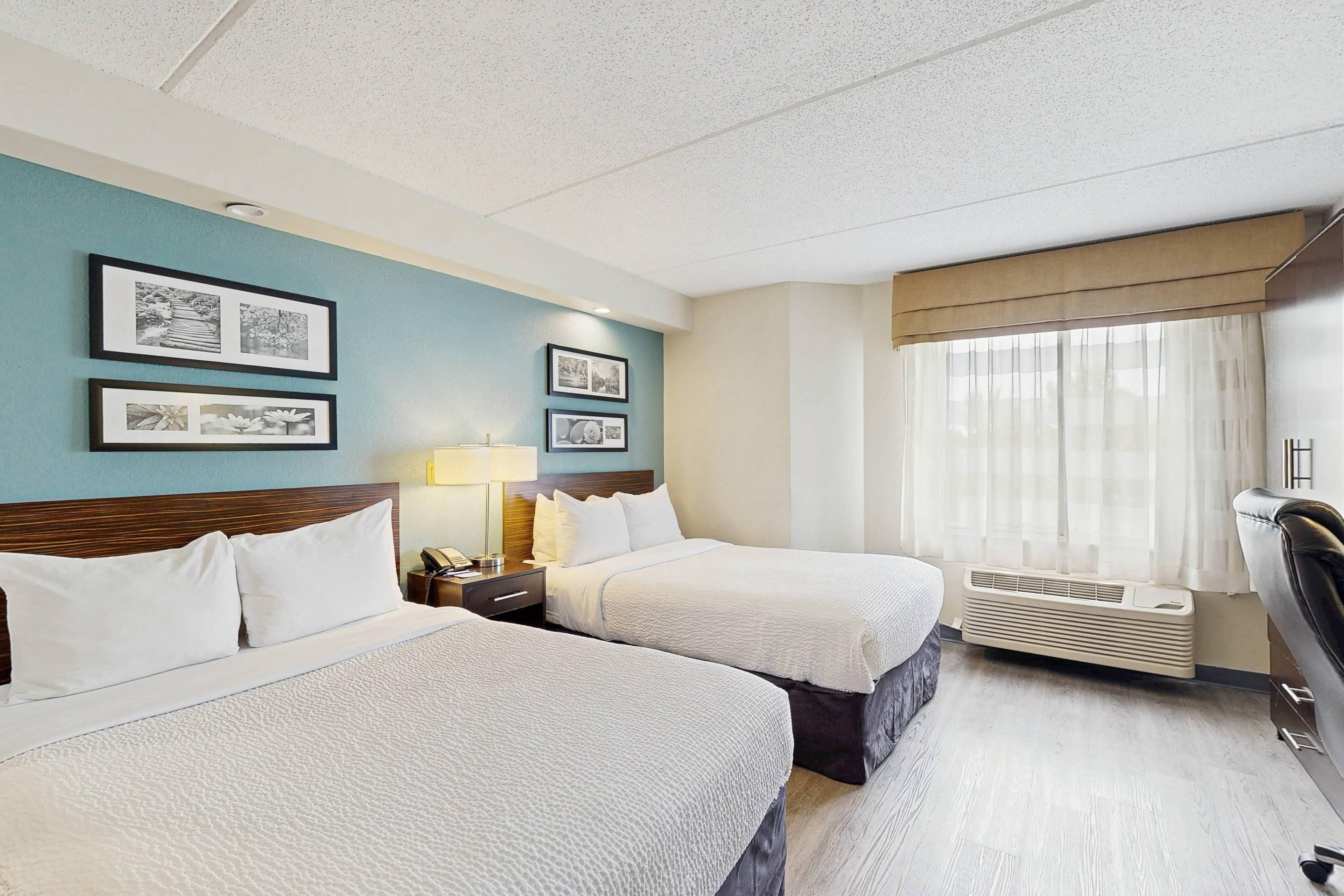 A hotel room with two beds, a desk with a lamp, a window with curtains, and framed pictures on the wall.
