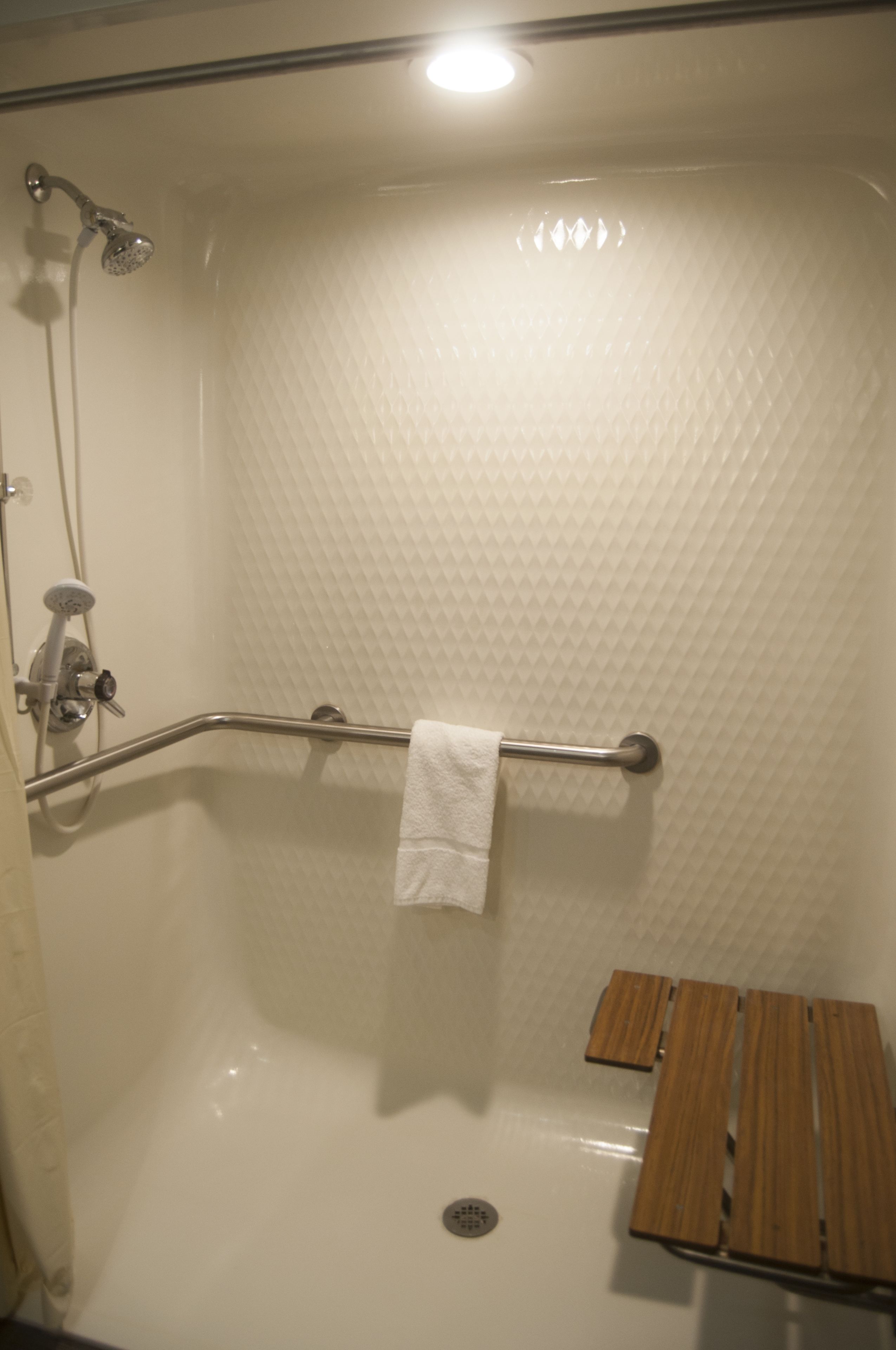 A walk-in shower with a white textured wall, a grab bar, a white towel, and a wooden bench.