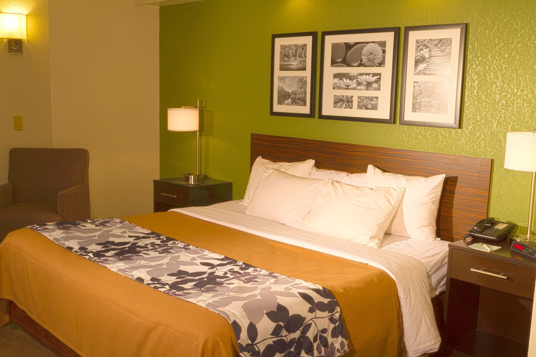 A hotel room with a bed, a chair, a nightstand with a lamp, and three framed pictures on the wall.