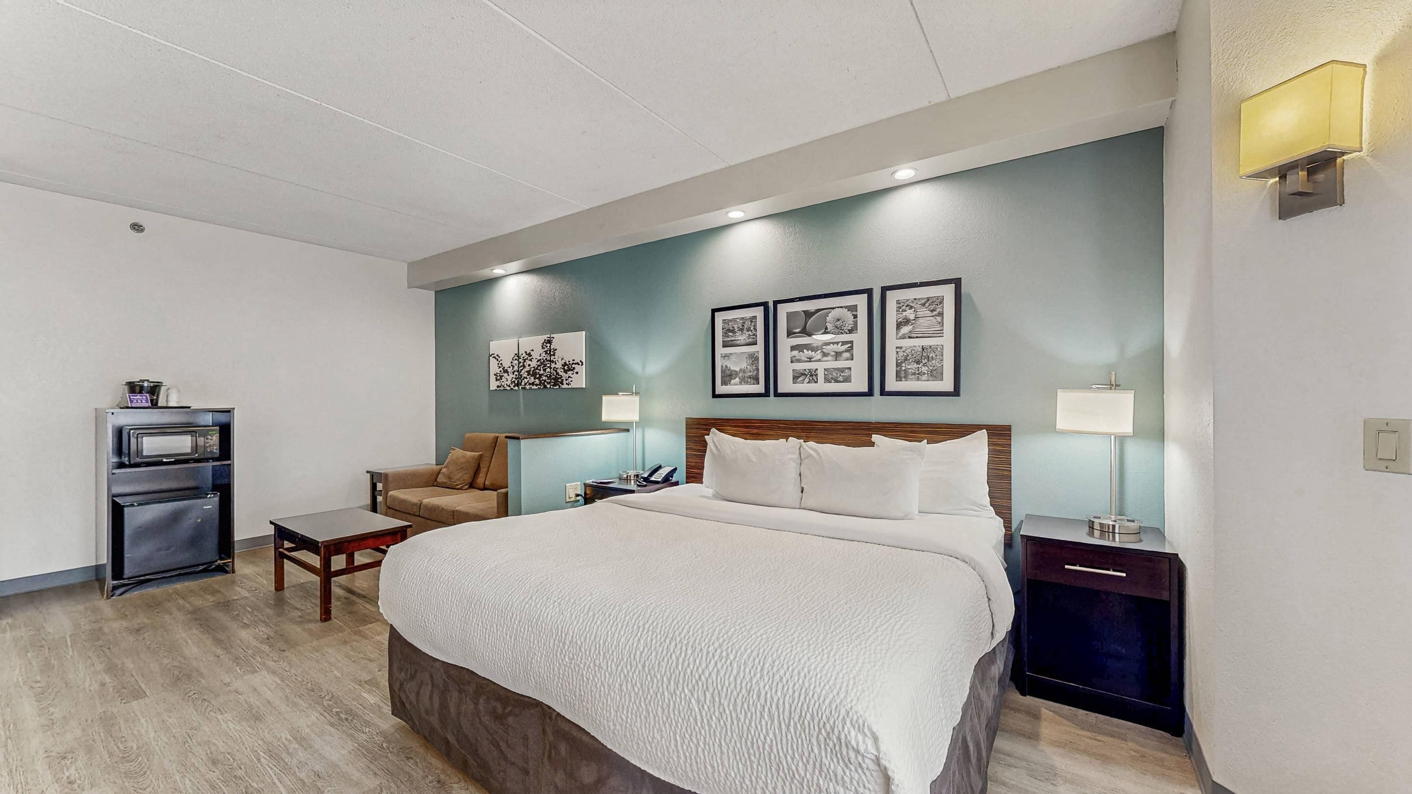 Una habitación de hotel con cama, zona de estar con una silla, una mesita, microondas y refrigerador.