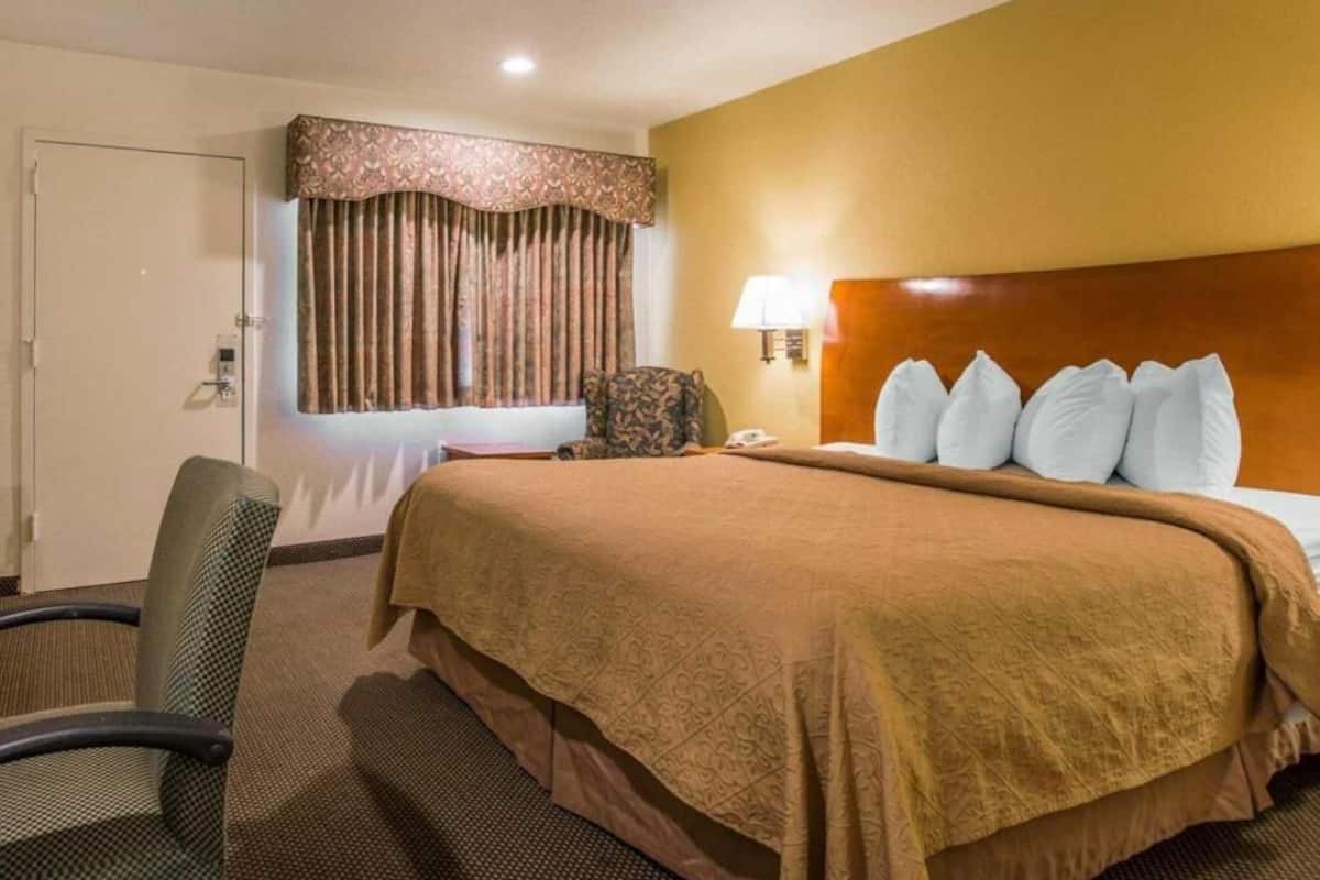 Una habitación de hotel con una cama, una silla, una lámpara y un ventanal con cortinas.