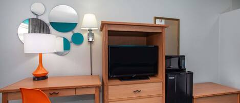 A compact hotel room with a wooden desk, a television, a refrigerator, a microwave, a chair, and wall-mounted mirrors.