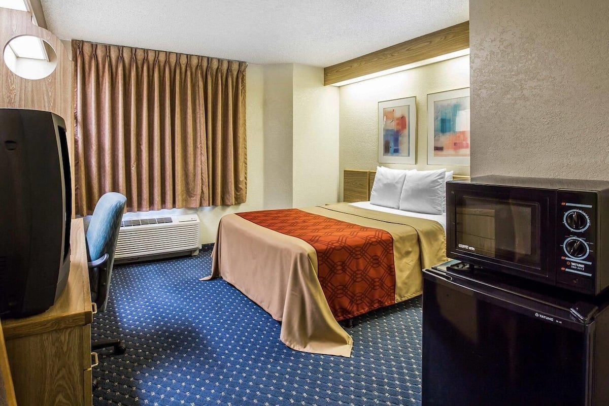 A hotel room with a bed, a desk with a computer, an air conditioning unit, a microwave, and a window with curtains.