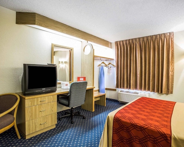 Habitación de hotel con escritorio de madera, televisor, silla, cama, espejo, armario y ventana con cortinas.