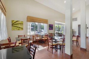 Un comedor con varias mesas y sillas, ventanas amplias con persianas y obras de arte en las paredes.