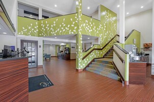 Un vestíbulo espacioso con un mostrador de recepción de madera, una escalera con diseños de hojas verdes y blancas, y una zona en el segundo piso con área de descanso y un mostrador de recepción.