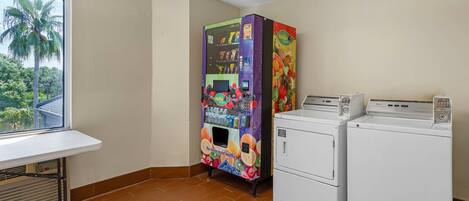 A laundry room with two washing machines, a vending machine, and a window with a view of palm trees.