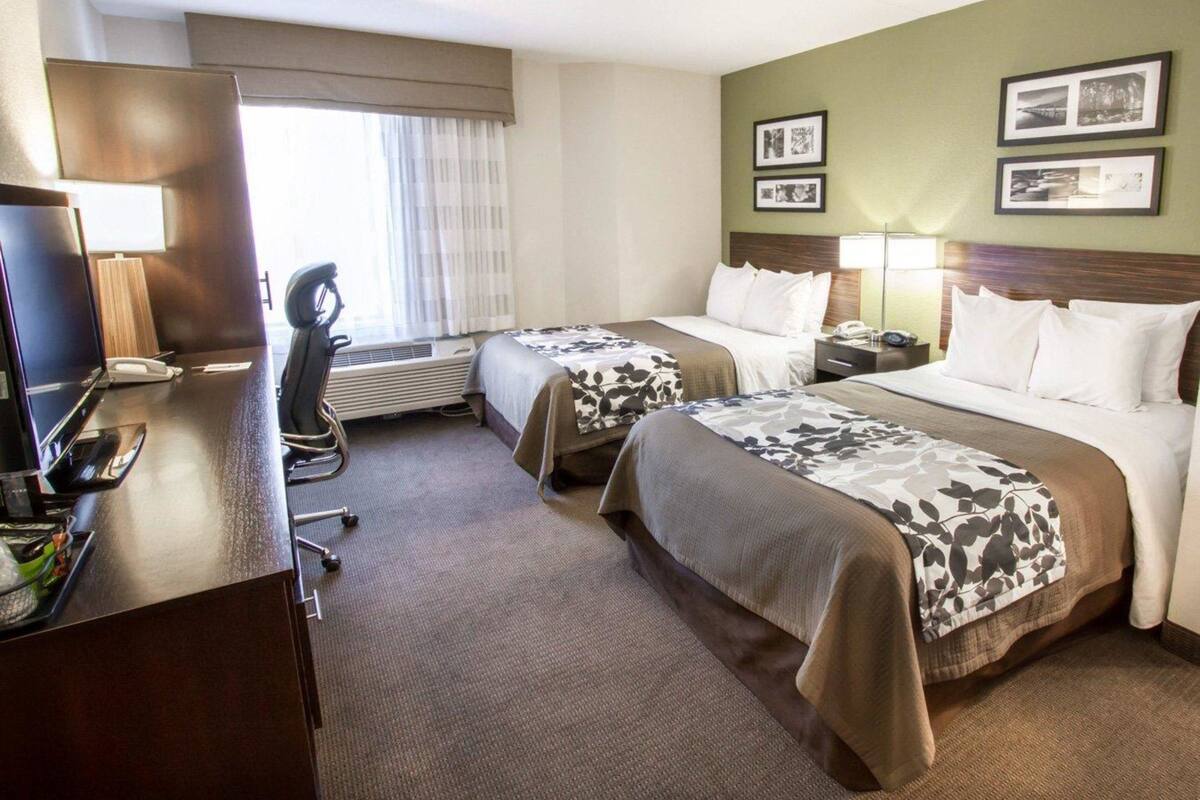 Habitación de hotel con dos camas, un escritorio con computadora y teléfono, una silla y una ventana con cortinas.