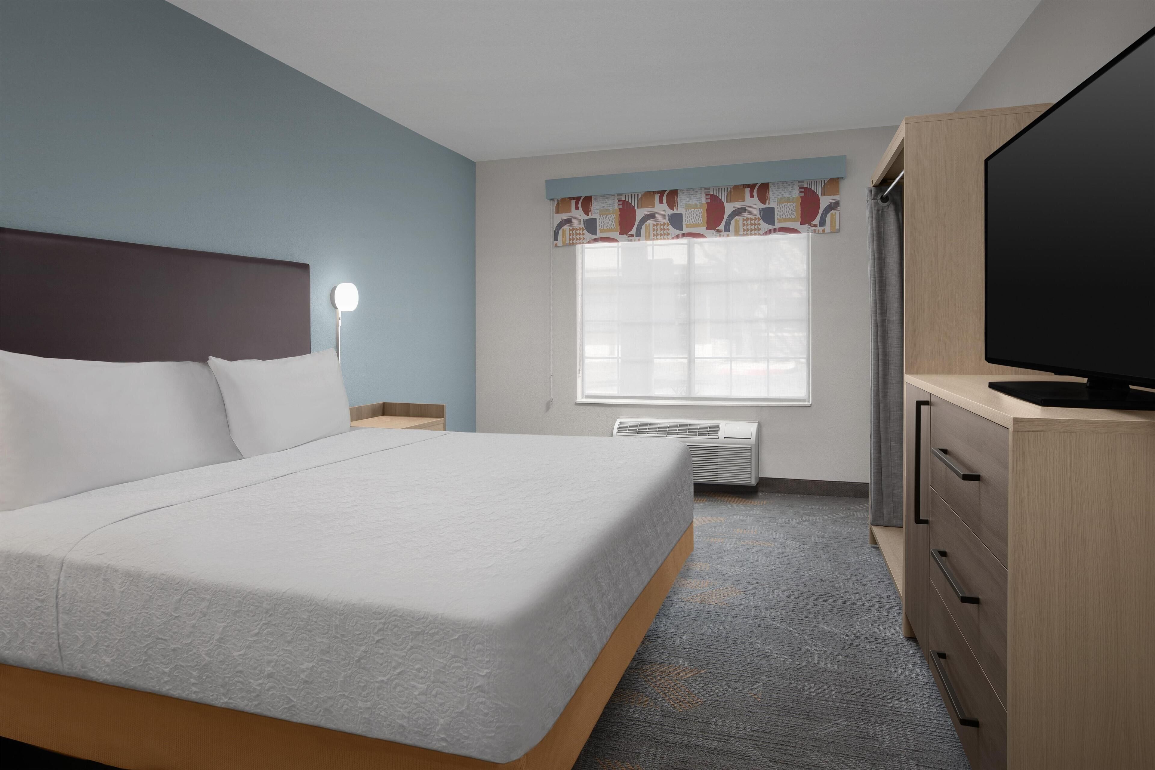 A hotel room with a bed, a television, a desk, and a window with curtains.