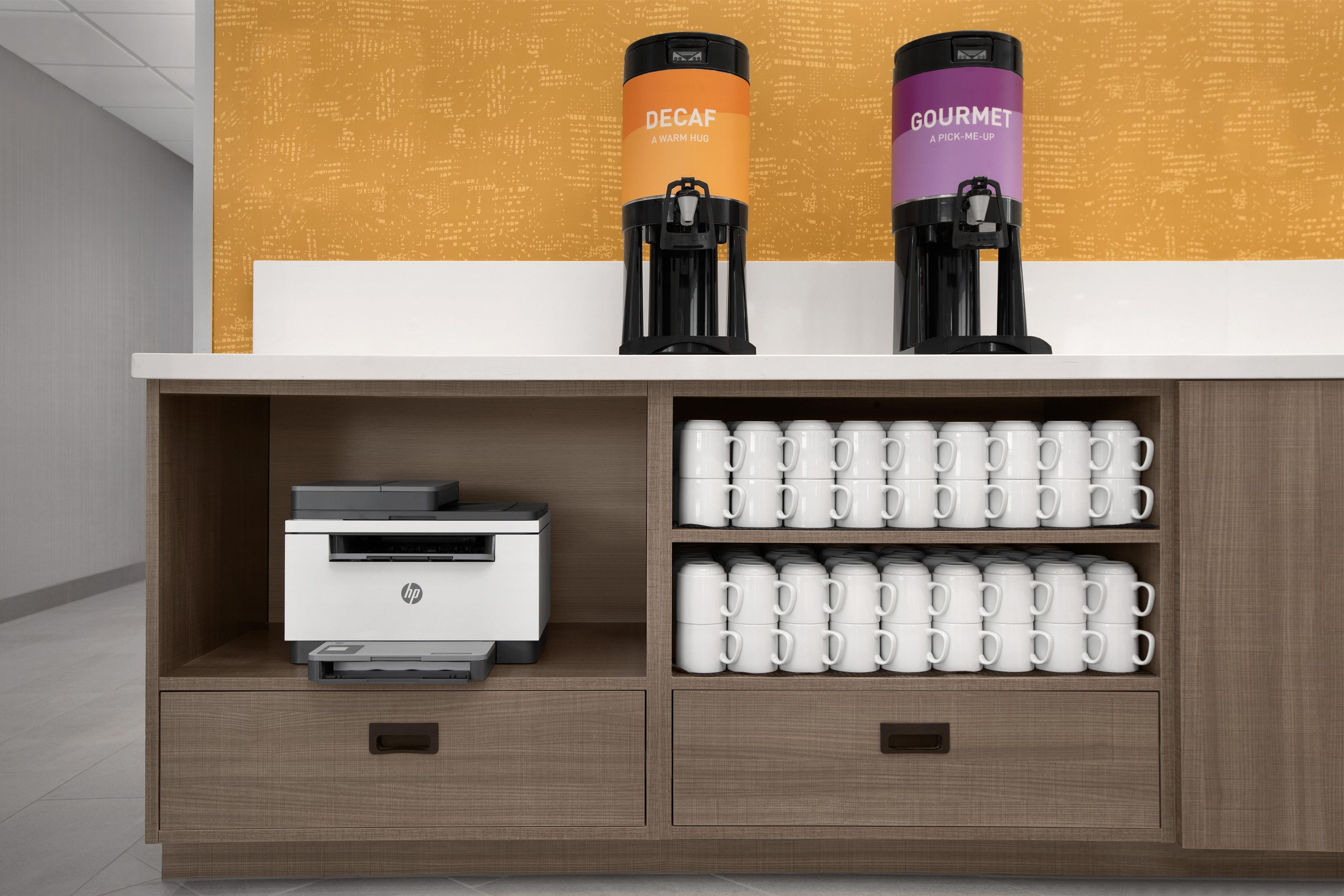 A coffee station with two coffee makers labeled "DECAF" and "GOURMET," a printer, and multiple white mugs on shelves.