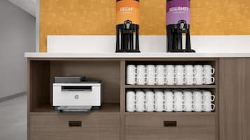 A coffee station with two coffee makers labeled "DECAF" and "GOURMET," a printer, and multiple white mugs on shelves.
