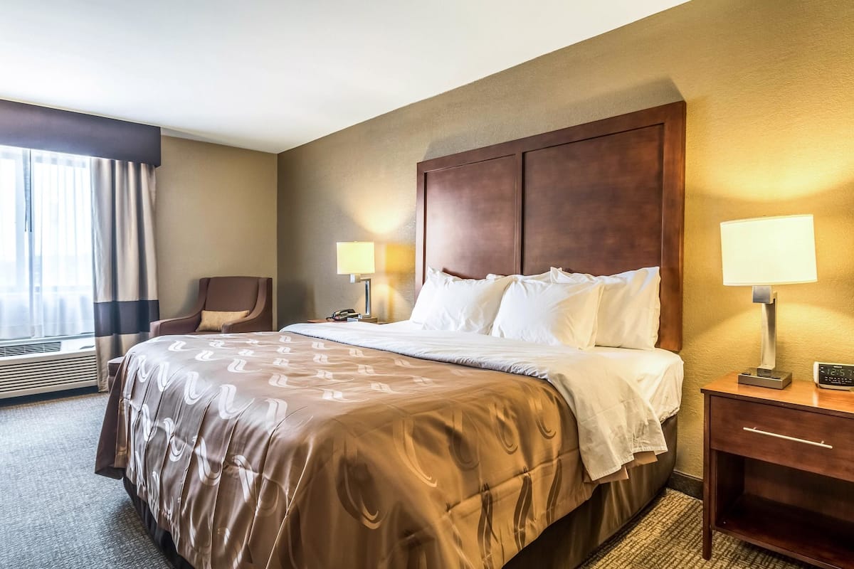 A hotel room with a large bed, two bedside lamps, a wooden nightstand, a chair, and a window with curtains.