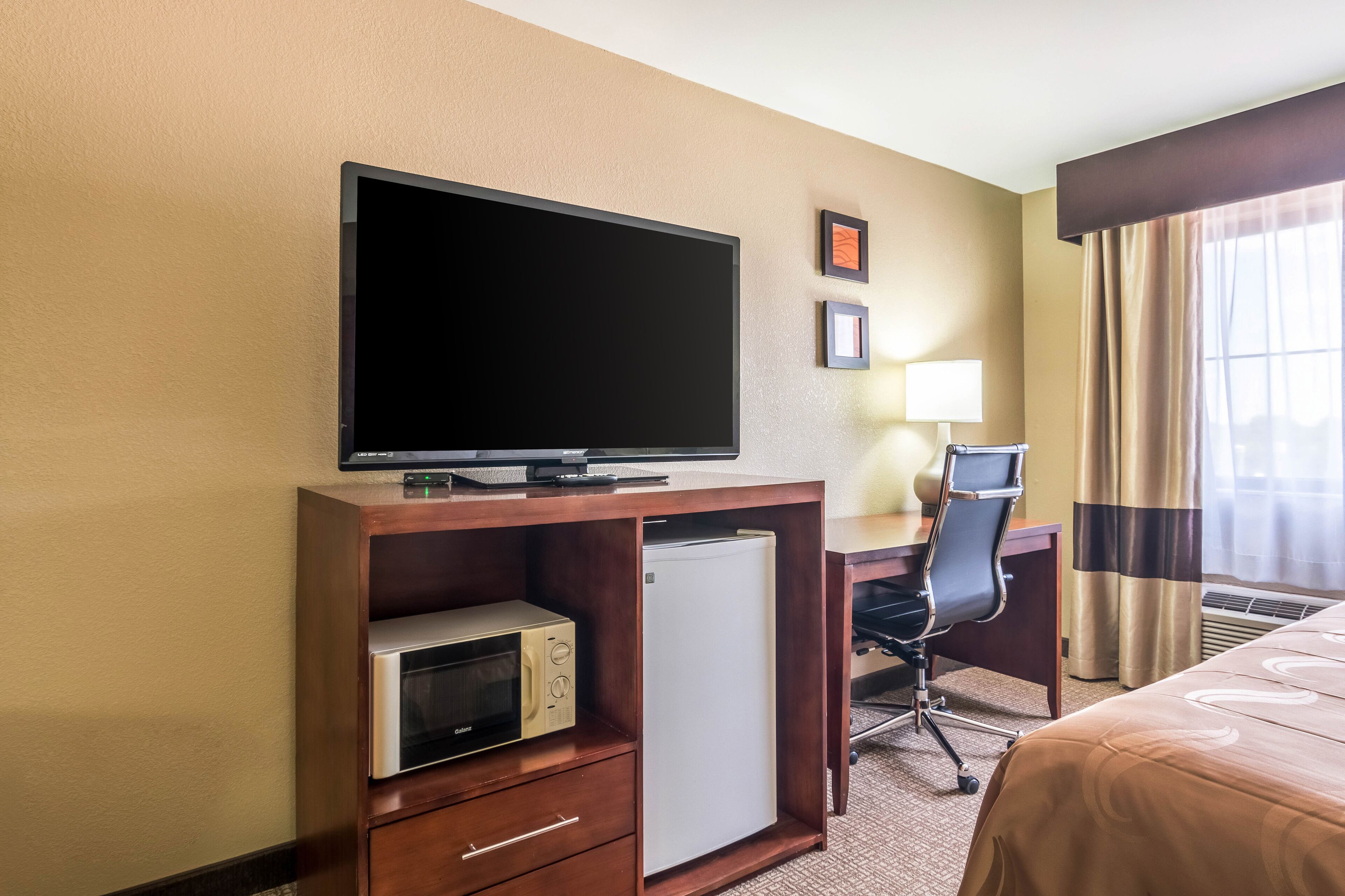 Quarto de hotel com televisão sobre suporte de madeira, micro-ondas, frigorífico, escrivaninha com cadeira, cama com colcha estampada e janela com cortinas.