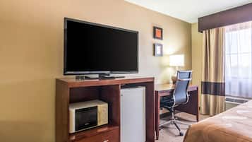 Quarto de hotel com televisão sobre suporte de madeira, micro-ondas, frigorífico, escrivaninha com cadeira, cama com colcha estampada e janela com cortinas.