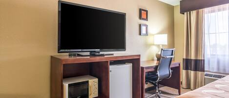 A hotel room with a television on a wooden stand, a microwave, a refrigerator, a desk with a chair, a bed with a patterned bedspread, and a window with curtains.