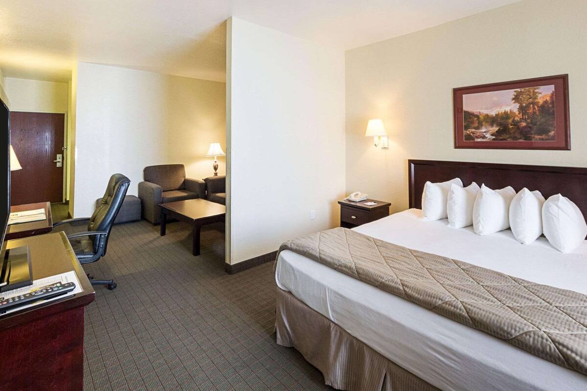 A hotel room with a bed, a desk with a computer, a chair, a small table, and a nightstand with a lamp.