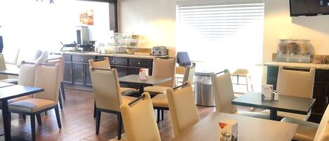 A restaurant with multiple tables and chairs, a kitchen area with a coffee machine, and a large window with blinds.
