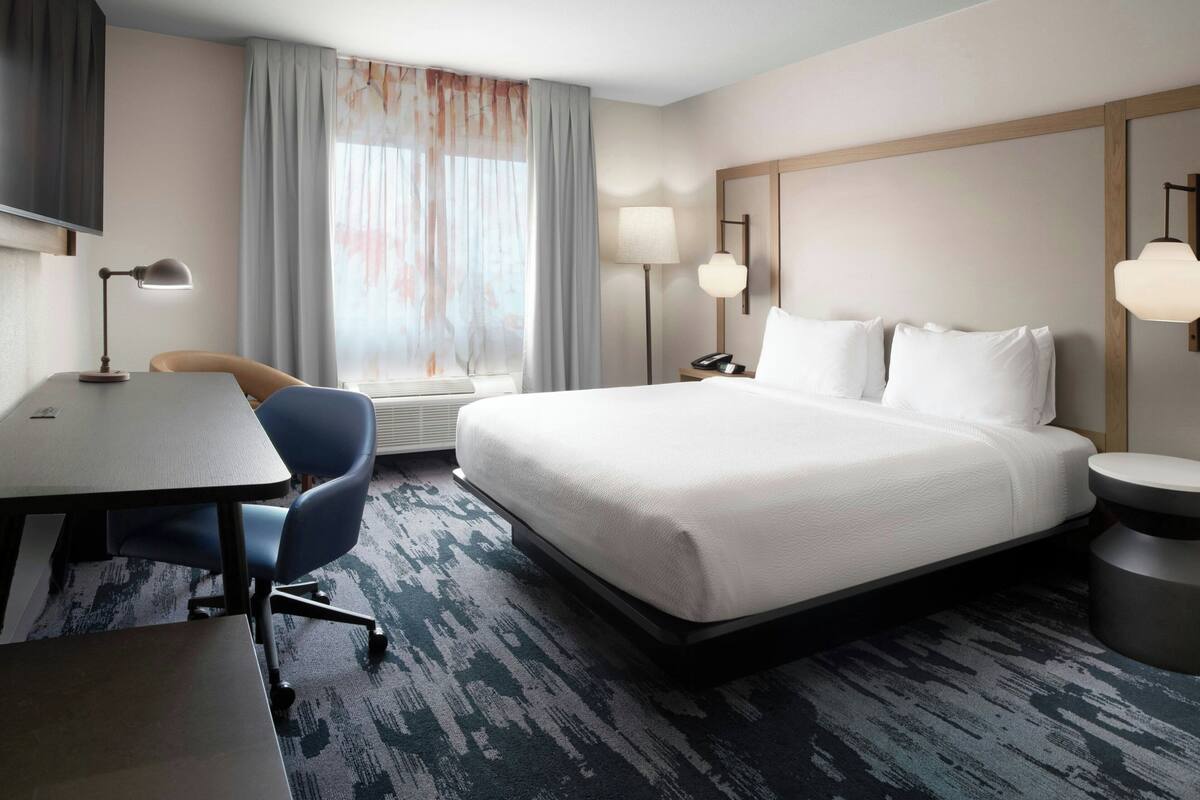 A hotel room with a bed, a desk, a chair, a lamp, and a window with curtains.