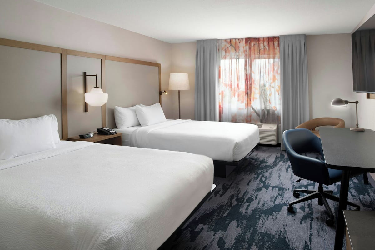 A hotel room with two beds, a desk, a chair, a lamp, and a window with curtains.