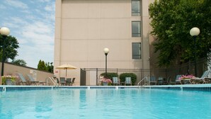 Une piscine entourée de chaises longues et de parasols, le tout bordé d’un bâtiment à la façade beige et d’une clôture.