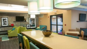 Un hall d’hôtel avec une longue table, des chaises vertes, un bol posé sur la table et un téléviseur accroché au mur.