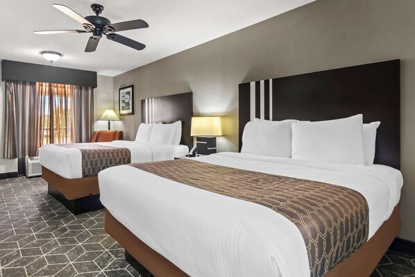 A hotel room with two beds, a ceiling fan, two bedside lamps, a large window with curtains, and a patterned floor.