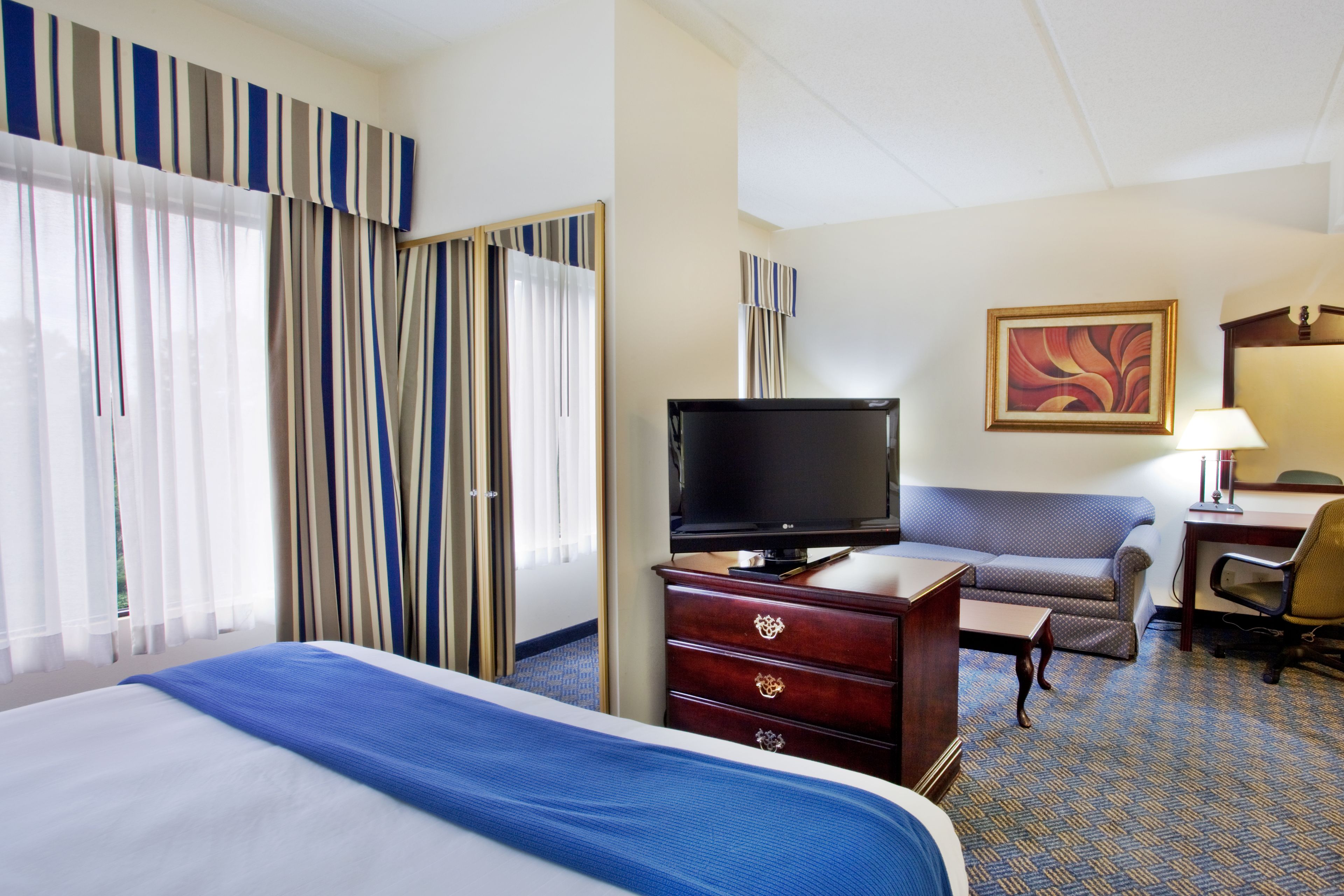 A hotel room with a bed, a TV on a dresser, a sofa, a desk with a lamp, and a painting on the wall.