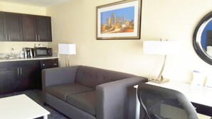 A hotel room with a gray sofa, a desk with a chair, a microwave, a coffee maker, a framed cityscape picture, and a lamp. - Holiday Inn Express Hotel & Suites by IHG (Lawrenceville)