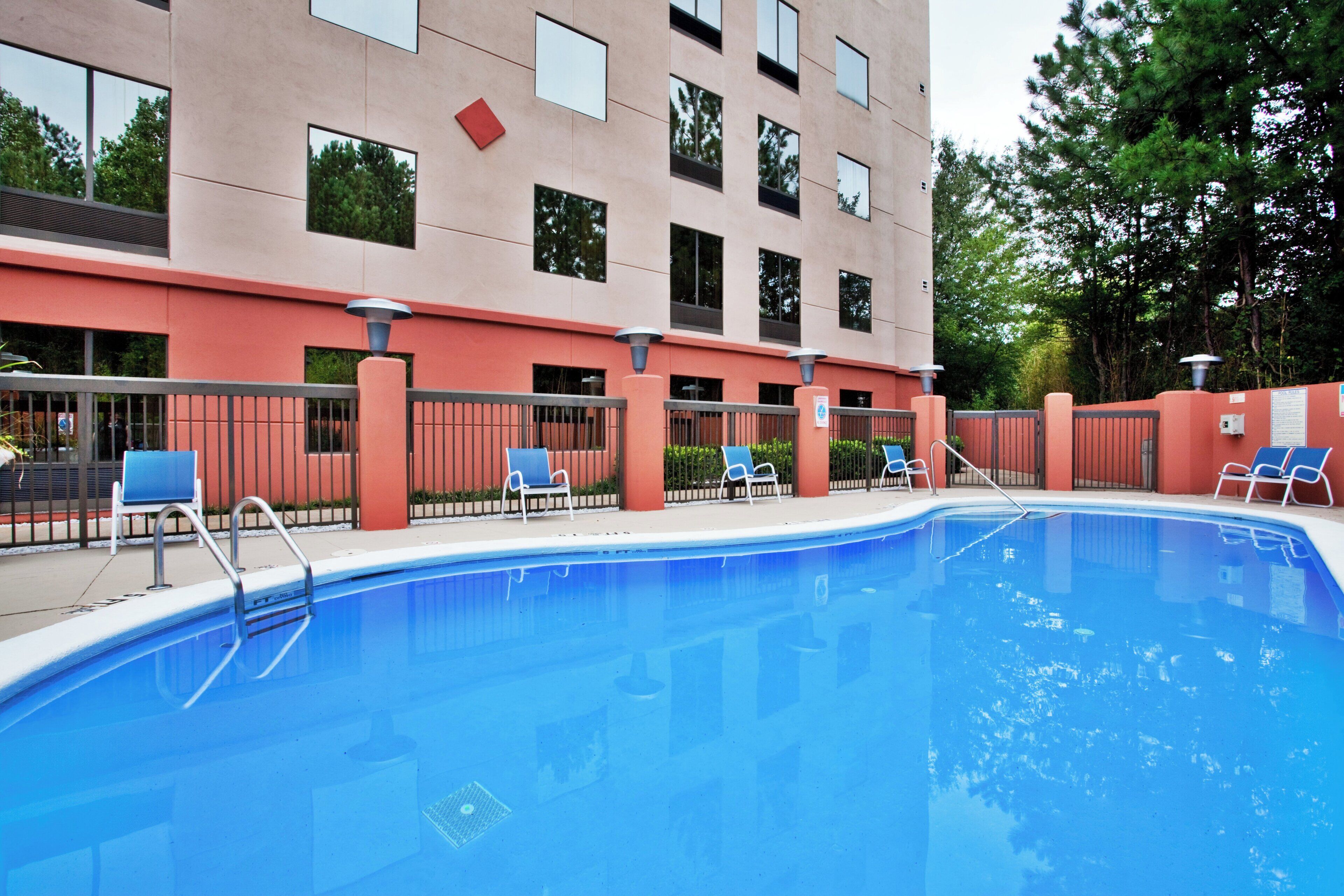 A swimming pool with blue water, surrounded by a fence, and a building with multiple windows and a red wall in the background.