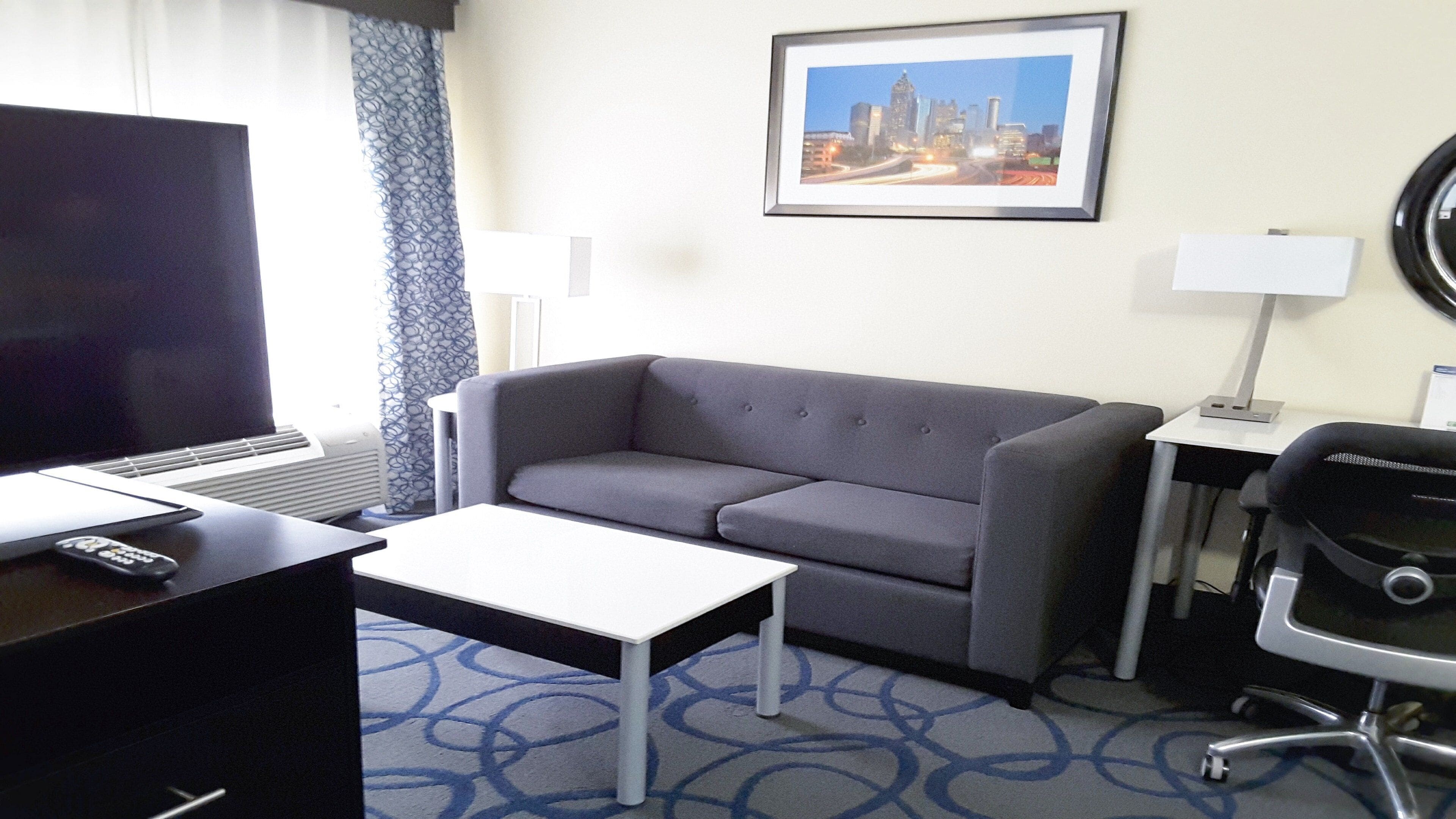 A hotel room with a grey sofa, a small white table, a TV, a desk with a lamp, and a framed cityscape picture on the wall.