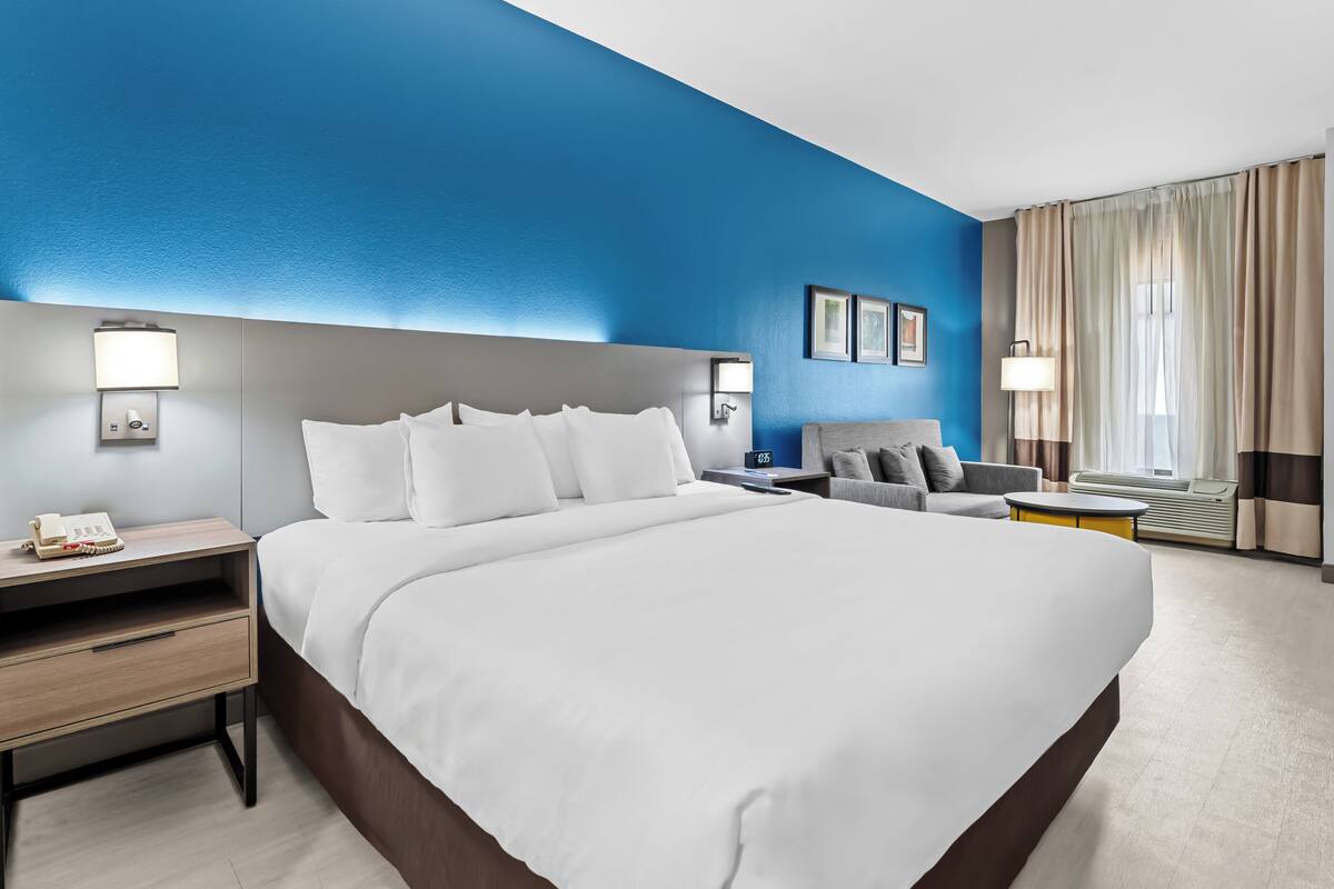 A hotel room with a large bed, a bedside table with a lamp, a chair, a small table, and a window with curtains.