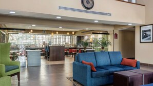 A hotel lobby with a blue sofa, green armchair, and a dark ottoman. There is a reception desk in the background, and a large clock on the wall. - Comfort Inn & Suites Perry National Fairgrounds Area (Perry)
