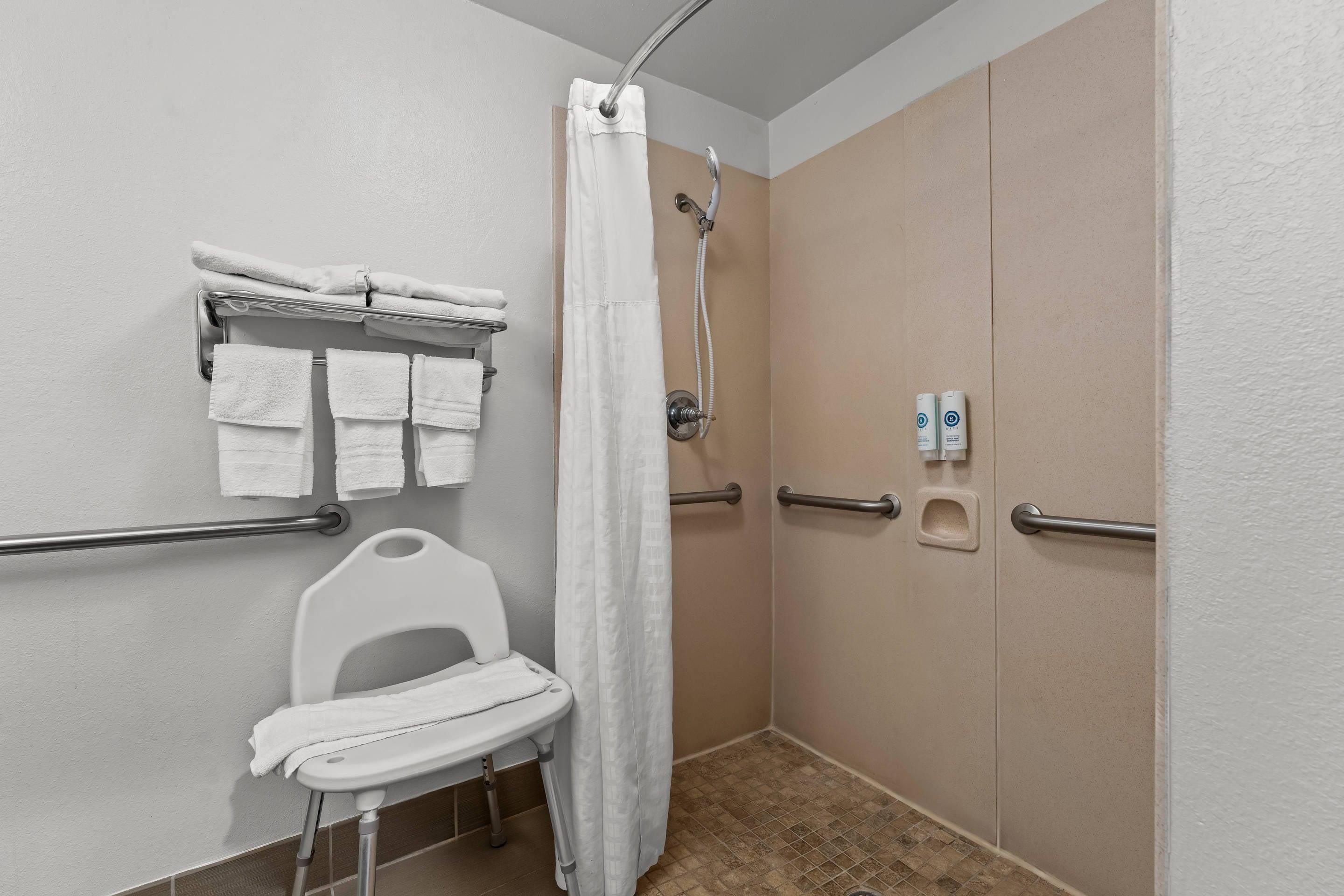 A bathroom with a shower, a grab bar, a white toilet with a seat, and a towel rack with multiple towels.