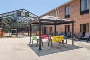 A covered outdoor seating area with chairs and a bench, a glass-enclosed building with a roof, and a brick building in the background. - Comfort Inn & Suites Perry National Fairgrounds Area (Perry)