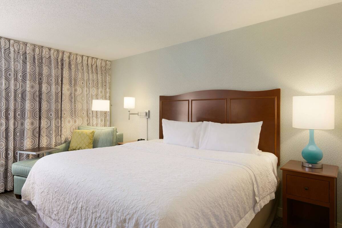 Ein Hotelzimmer mit einem großen Bett, einem Nachttisch mit Lampe, einem Sessel mit Kissen und gemusterten Vorhängen.