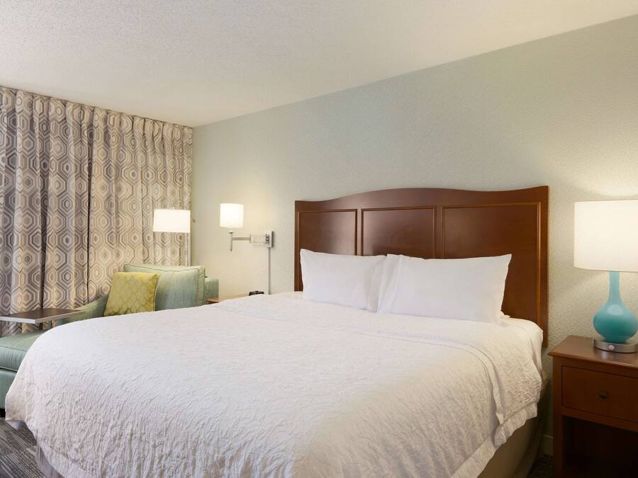 Quarto de hotel com uma cama grande, mesa de cabeceira com abajur, cadeira com almofada e cortinas estampadas.