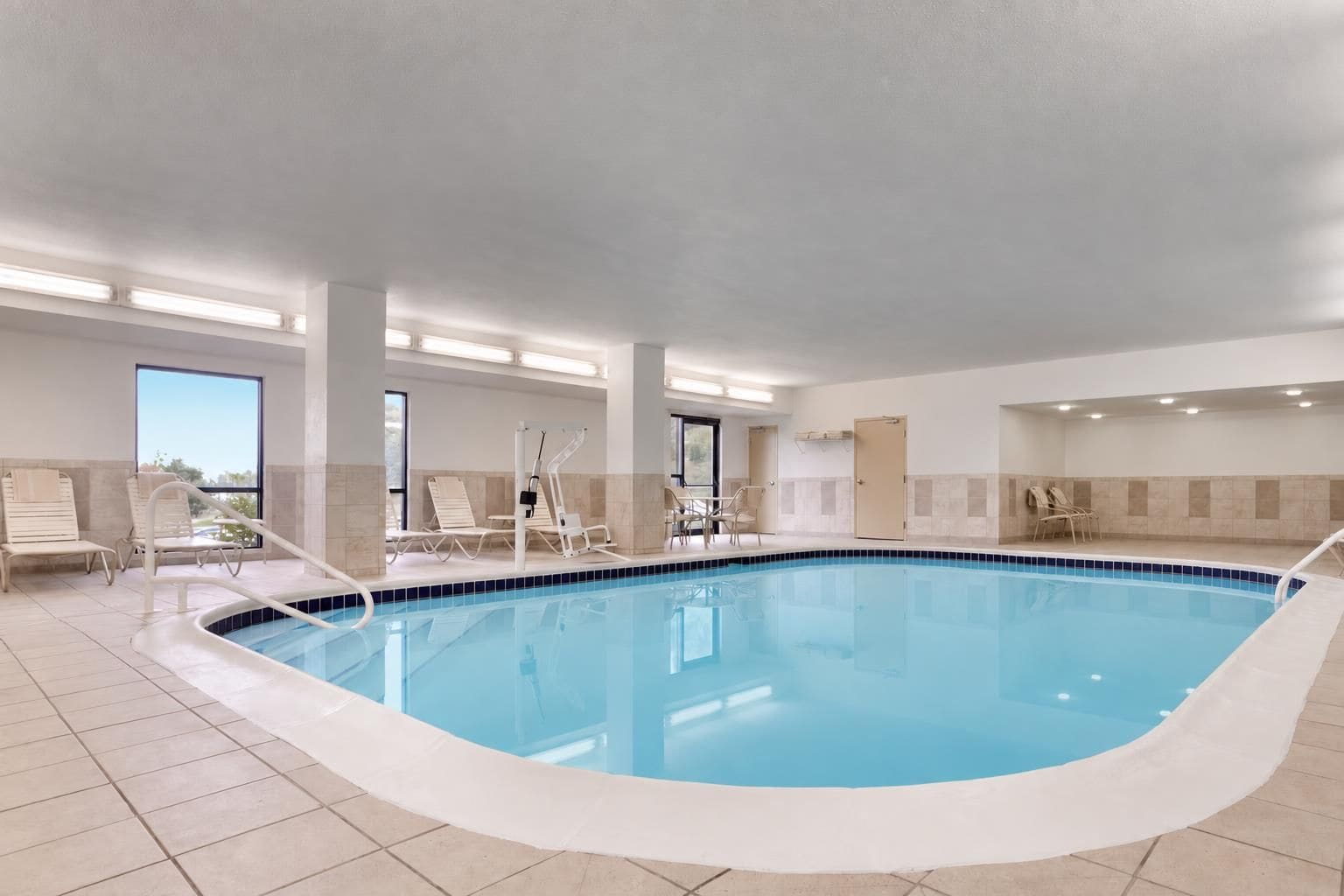 Una piscina interior con suelo de baldosas, sillas blancas junto a la piscina y una vista despejada del cielo a través de amplias ventanas.