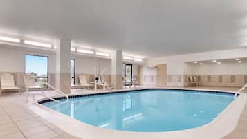 Una piscina interior con suelo de baldosas, sillas blancas junto a la piscina y una vista despejada del cielo a través de amplias ventanas.